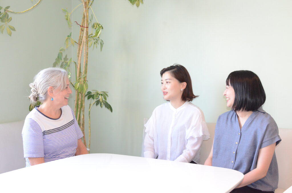 Three women of different ages sit around a white table chatting in a light green room with a tall plant nearby.